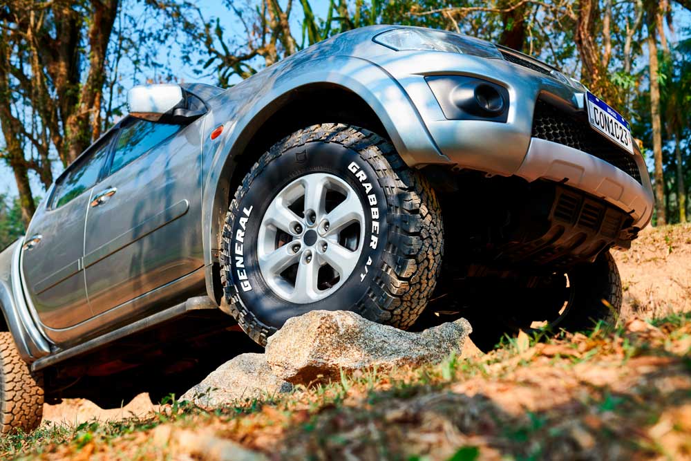 Imagem - Notícia sobre Chegada do Grabber A/Tx no Brasil Um carro off-road cinza está subindo uma rocha em um ambiente natural com árvores ao fundo. O veículo está equipado com pneus General Tire Grabber A/Tx, projetados para terrenos mistos.
