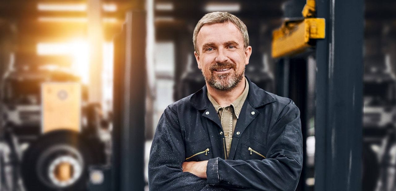 A smiling garage owner stands looking to camera from his busy auto repair shop .