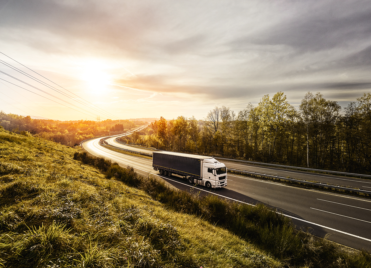 White truck driving on the highway turning towards the horizon in an autumn landscape with sun shining through the clouds in the sky