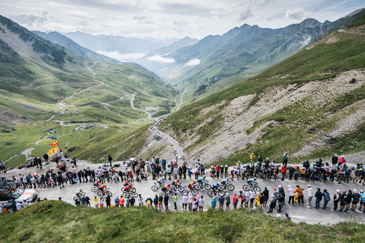 Cyclists climb a winding mountain road in the Pyrenees, cheered on by crowds against a backdrop of dramatic green valleys and rugged peaks.