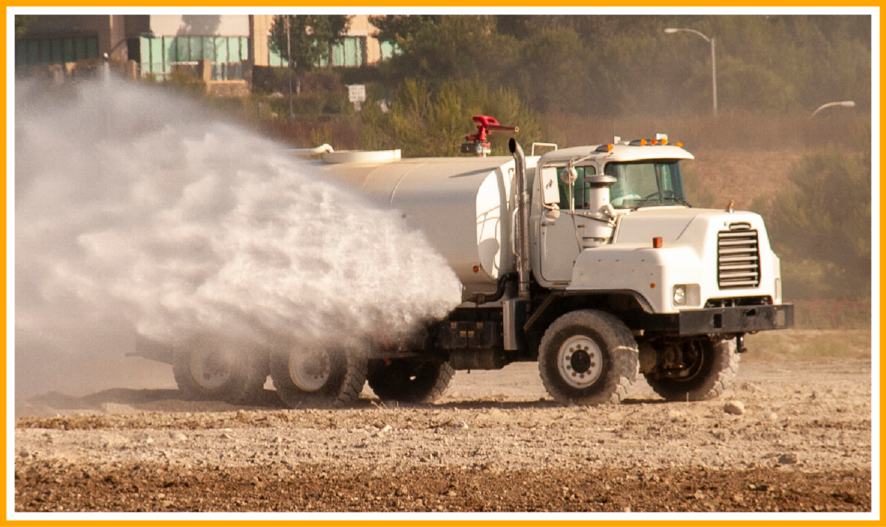White water truck spraying mist onto dry ground, with trees and buildings in the background