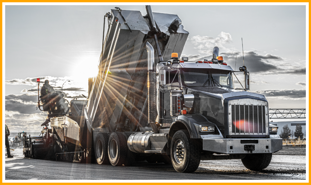 Dump truck unloading asphalt into a paving machine on a sunny road construction site.